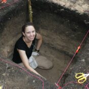 Archaeologist Amanda Finley in a 1 meter by 1 meter pit in Fiji. She is wearing a black shirt, kneeling at the bottom of the pit making a map of the unit.