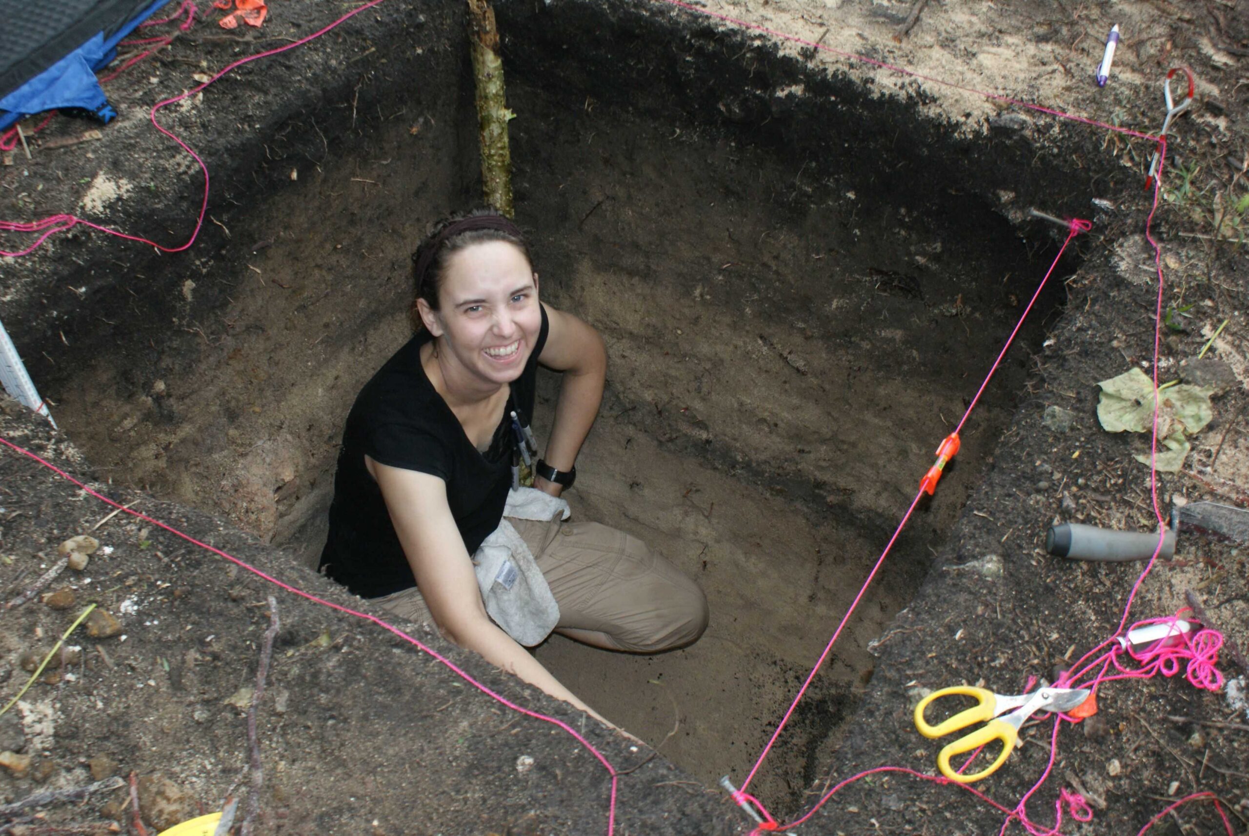 Archaeologist Amanda Finley in a 1 meter by 1 meter pit in Fiji. She is wearing a black shirt, kneeling at the bottom of the pit making a map of the unit.